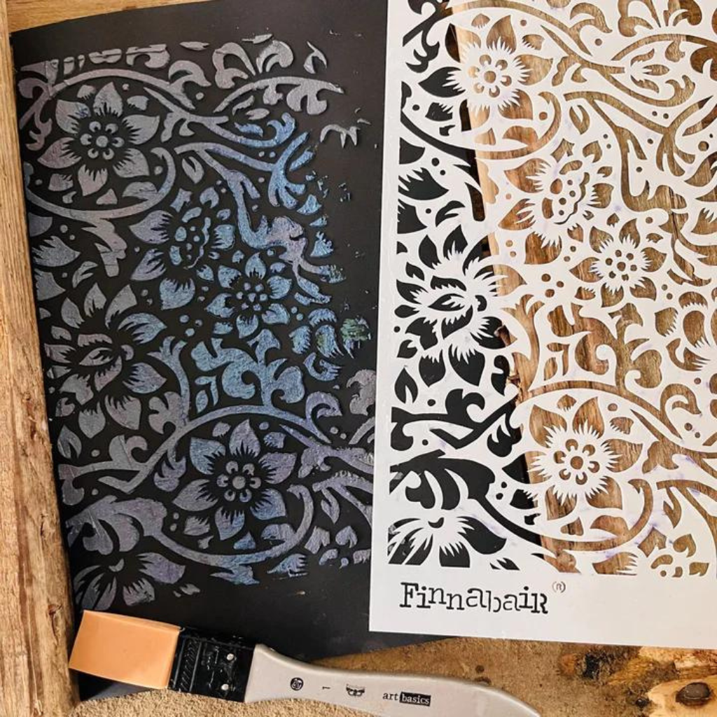 White Finnabair stencil with lace design. Next to it, a black canvas with white stenciling and paint brush.