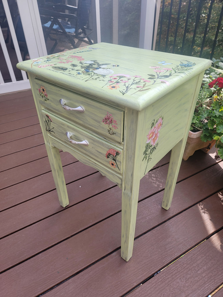 Upcycled green nightstand with floral rub-on transfers in a garden setting, showcasing DIY furniture makeover.