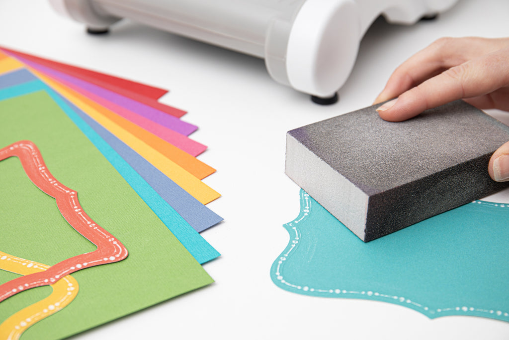 Person using a sanding block on a piece of paper with colorful sheets and a white machine in the background.
