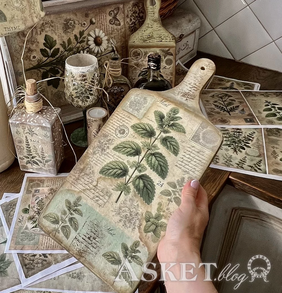 Decorative cutting board with botanical design held by a hand, surrounded by similar items on a wooden surface.