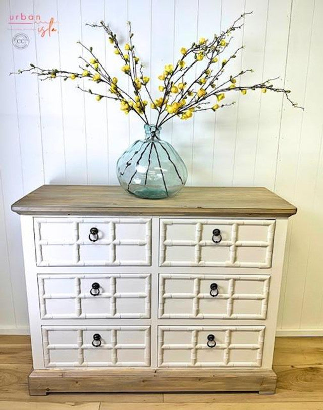 White dresser with wooden top and vase of yellow flowers against a white wall.