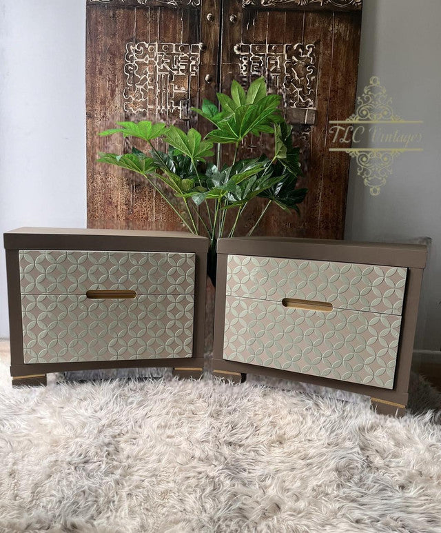 Two wooden storage boxes with decorative patterns on a textured surface, with a plant and wooden panel in the background.