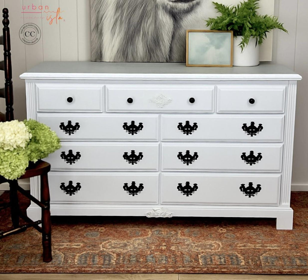White dresser with black decorative handles in a room setting with plants and artwork.