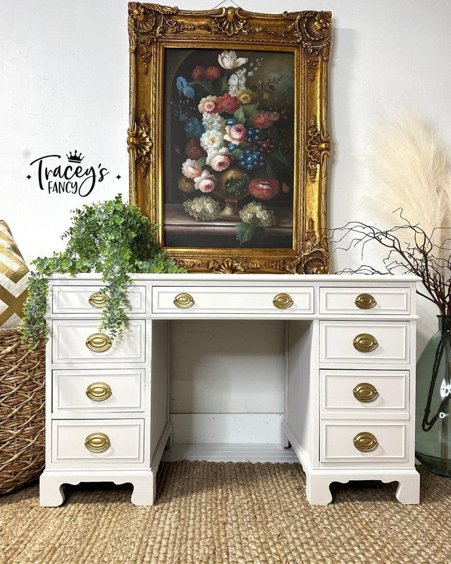 White desk with gold accents in a room with a decorative painting and plants.