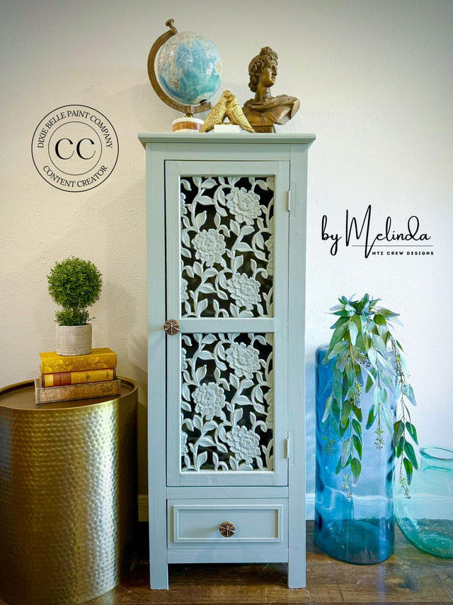 Decorative cabinet with floral patterns on a wooden floor, surrounded by plants and decor items.
